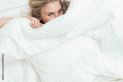 Happy Caucasian Blonde Girl Peeking from Under White Duvet Covers