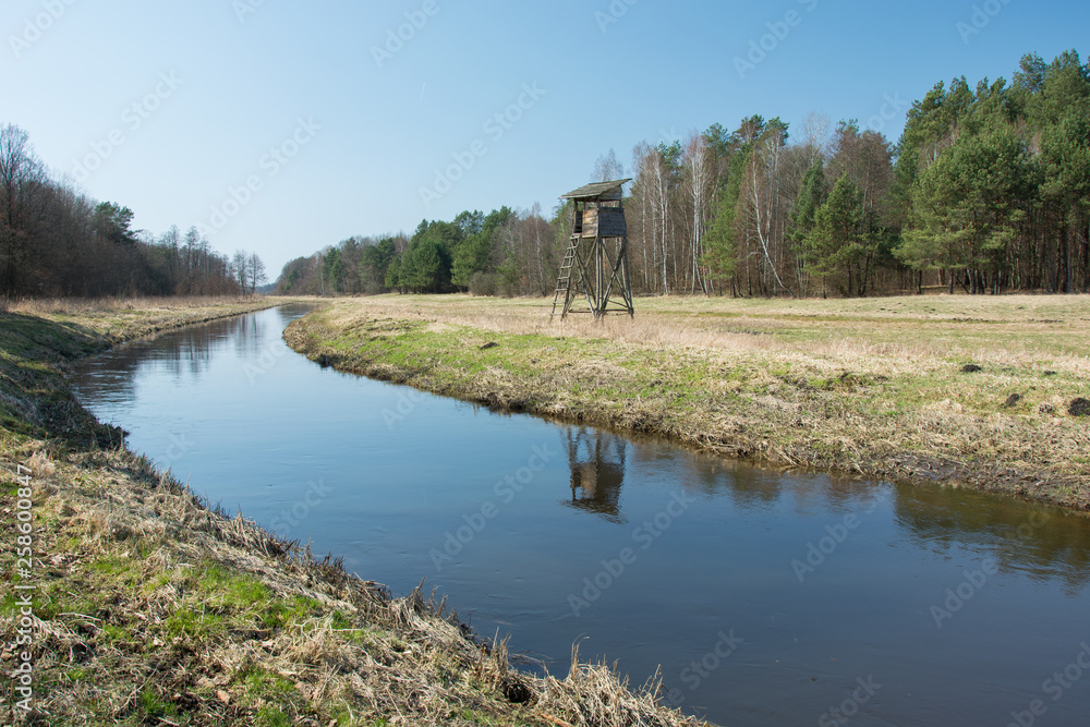 Quiet river flowing through a meadow, a hunting pulpit in the forest and blue sky