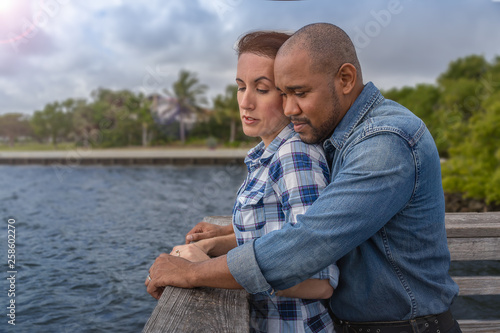  A multiracial couple looks over the pier. He embraces her from behind as they look over the pier on a windy day have a romantic moment.