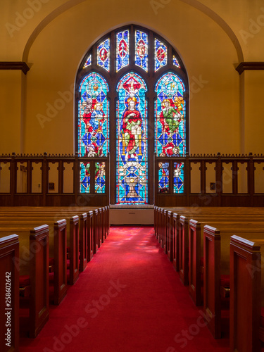 Red carpeted church aisle between pews illuminated by light from stained glass window