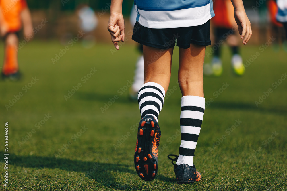 Fototapeta premium Legs of boy footballer in boots football cleats. Player walking on green grass soccer field at the stadium. Close-up of football sports equipment