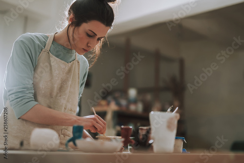 Beautiful young woman in apron painting pottery in workshop