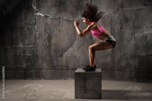 Fit young woman box jumping at a crossfit style on gray background. Fitness, crossfit, functional, training, and lifestyle concept
