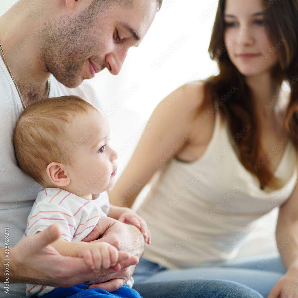 Loving parents sitting in living room at home affectionately cradling adorable baby son in their arms looking at him. Childhood, parenthood, togetherness, love, happiness concept Loving parents sitting in living room at home affectionately cradling adorable baby son in their arms looking at him. Childhood, parenthood, togetherness, love, happiness concept