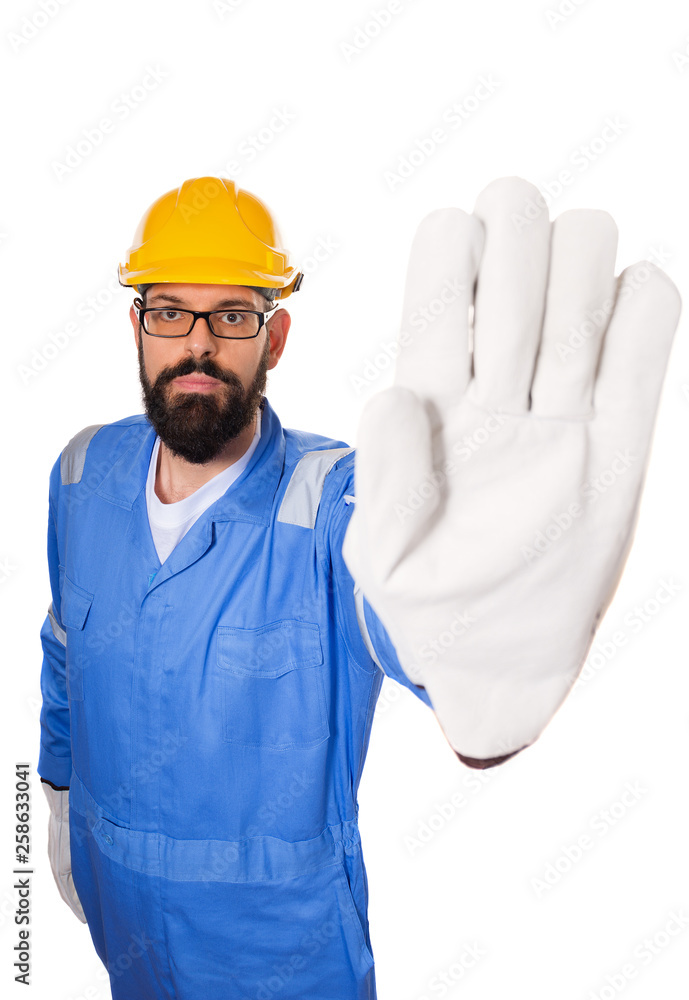 Fun high angle of bearded workman with yellow helmet, blue uniform and in glasses making stop gesture with his hand in gloves. over white background Fun high angle of bearded workman with yellow helmet, blue uniform and in glasses making stop gesture with his hand in gloves. over white background