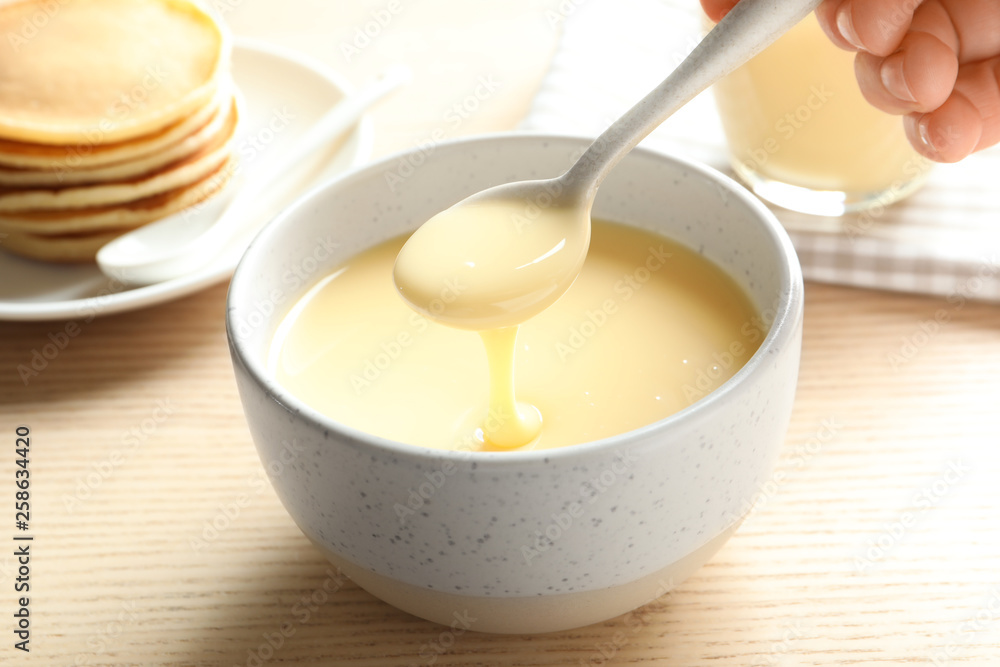Person holding spoon with condensed milk over bowl on table, closeup. Dairy products Person holding spoon with condensed milk over bowl on table, closeup. Dairy products
