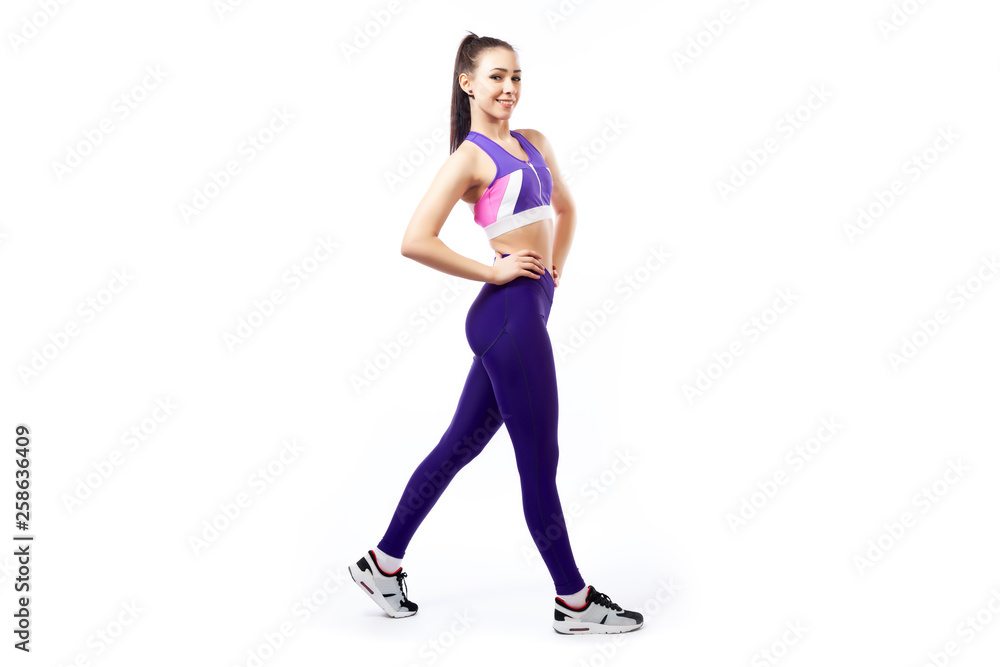 A young woman coach in a sporty piurple short top and gym leggings posing, doing exercise, step forward on a white isolated background in studio A young woman coach in a sporty piurple short top and gym leggings posing, doing exercise, step forward on a white isolated background in studio