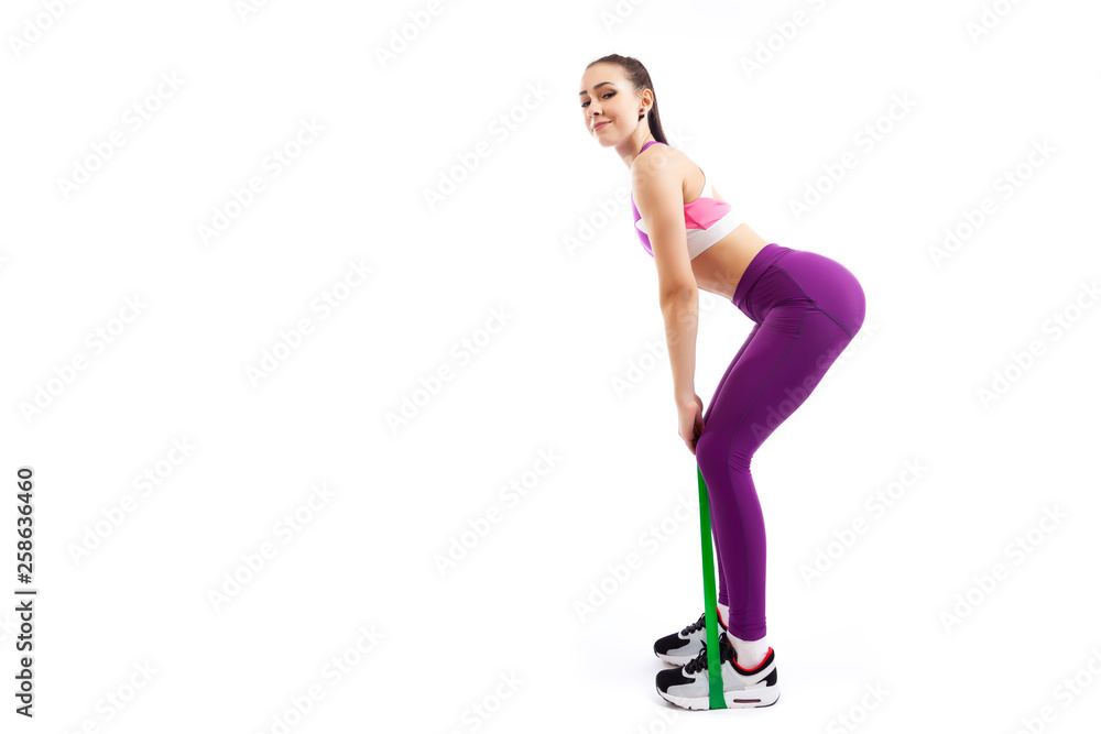 A young woman coach in a sporty pink short top and gym shows the correct squatting technique with sport fitness rubber bands; the position of a full squat on a white isolated background in studio A young woman coach in a sporty pink short top and gym shows the correct squatting technique with sport fitness rubber bands; the position of a full squat on a white isolated background in studio