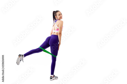 A ypung woman coach in a sporty purple short top and gym leggings makes lunges by the feet forward with sport fitness rubber bands, stretch legs on a white isolated background in studio