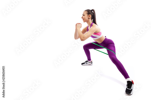 A young woman coach in a sporty pink short top and gym shows the correct squatting technique with sport fitness rubber bands; the position of a full squat on a white isolated background in studio