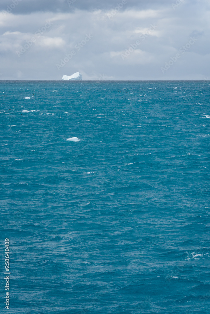 Restless nature background of choppy glacial melt water, floating ice chunks, stormy sky, and iceberg in background