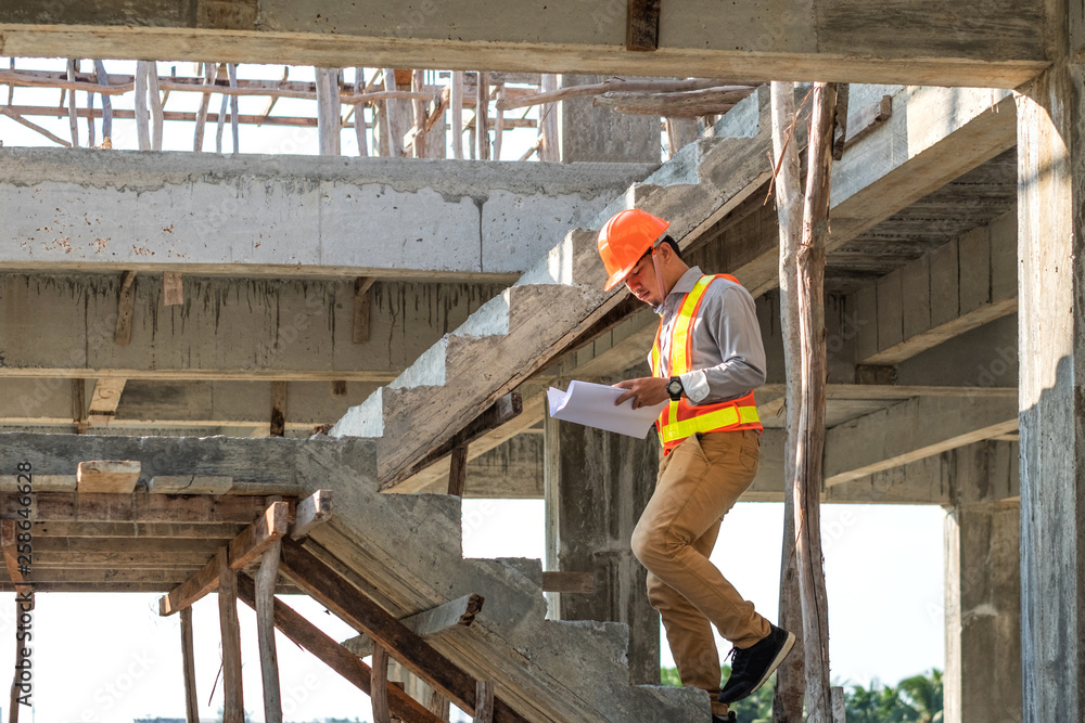 Asian architect or engineer holding looking project blueprint papers and wear helmet at construction site. He walking on stairs. Asian architect or engineer holding looking project blueprint papers and wear helmet at construction site. He walking on stairs.