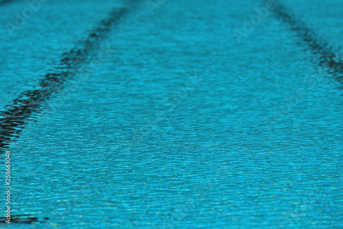 A swimming pool with a sunny reflection and beautiful wavy water surface in turquoise blue color with grab bars ladder on the sides and black line lanes underwater.