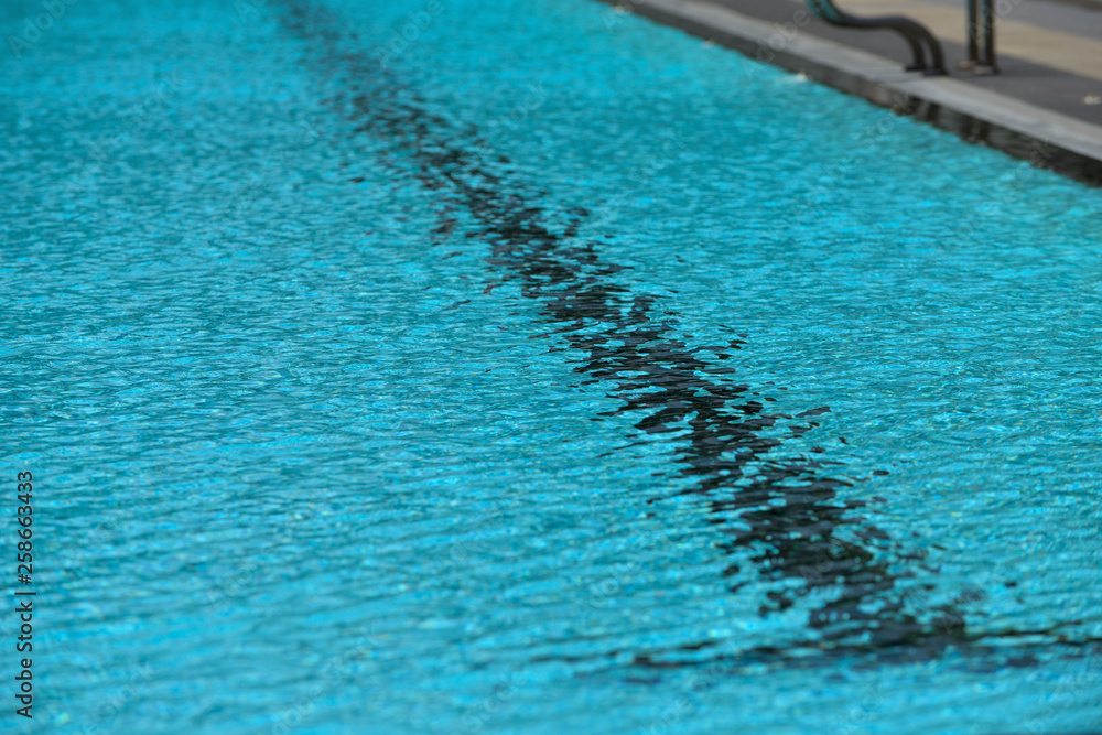 A swimming pool with a sunny reflection and beautiful wavy water surface in turquoise blue color with grab bars ladder on the sides and black line lanes underwater.