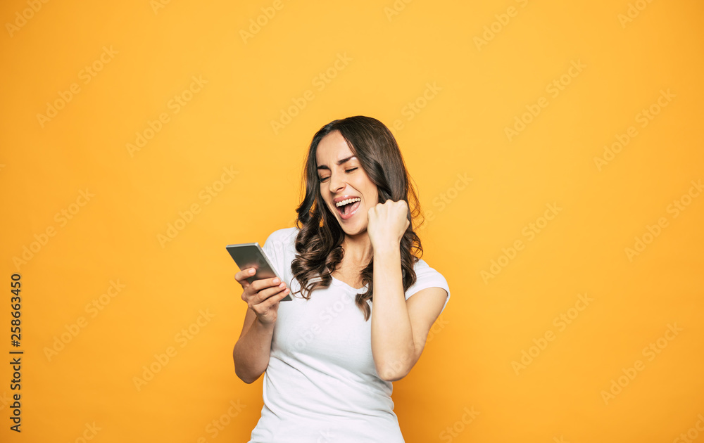 Hell yes! Screaming from good news girl with nice hairstyle and perfect skin in front of spicy-orange color is holding her phone and smiling sincerely. Hell yes! Screaming from good news girl with nice hairstyle and perfect skin in front of spicy-orange color is holding her phone and smiling sincerely.