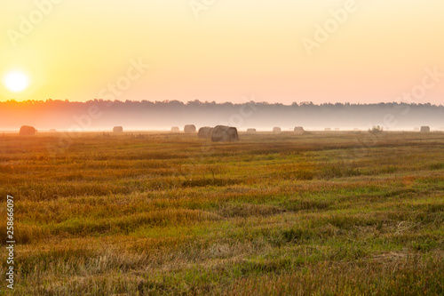 Rising sun dispersing morning mist in the field with bales of hay