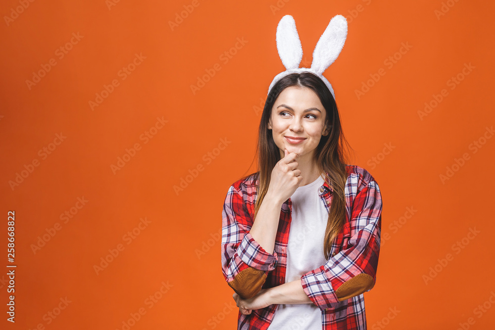 Beautiful young brunette woman in bunny ears looking at camera while standing against orange background.