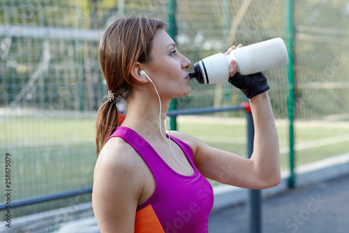 Attractive young caucasian woman in bright sportswear drinks water during fitness on outdoors sportsground. Active lifestyle woman hydrating with water sports bottle. Copy space.