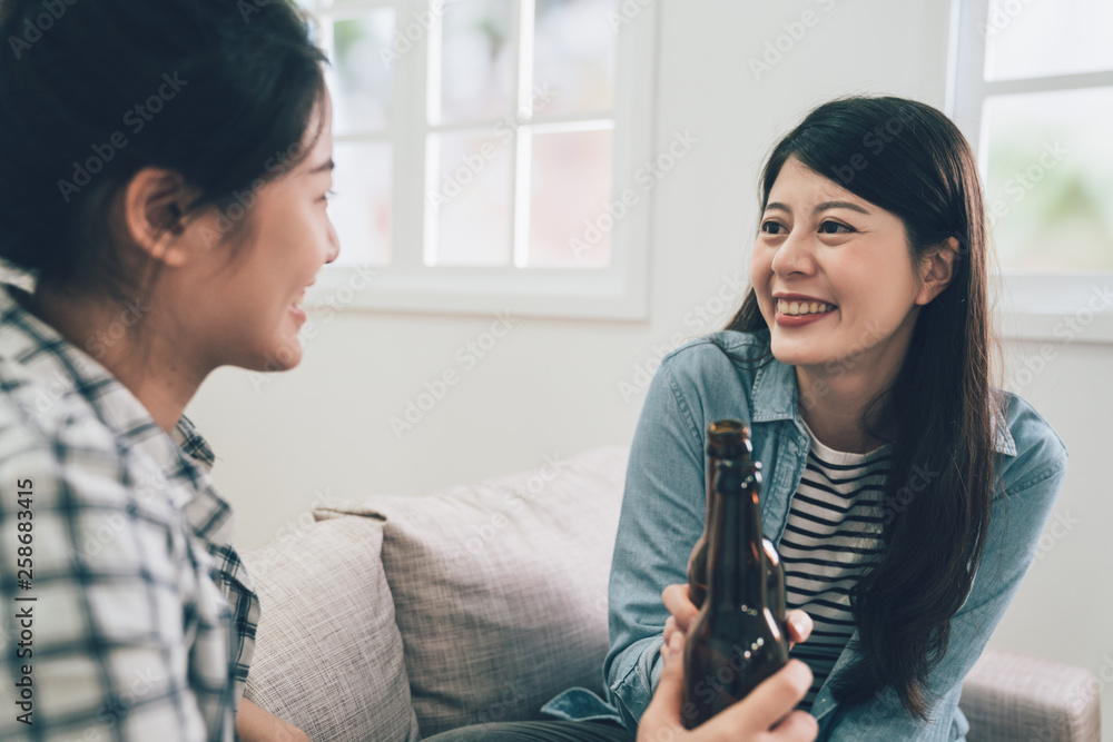 Fototapeta premium two asian female friends drinking beers together sitting on comfort couch sofa in living room in dormitory. girl roommates looking each other cheers making toast together celebrating new relationship