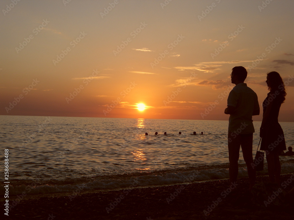 Silhouette of a guy and a girl watching the sun go beyond the sea horizon. Sunset in red tones. Sky, water, men and women. Silhouette of a guy and a girl watching the sun go beyond the sea horizon. Sunset in red tones. Sky, water, men and women.