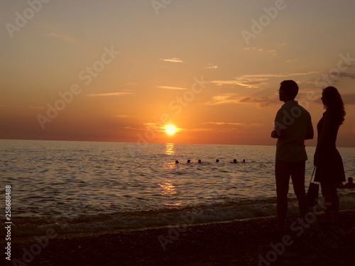 Silhouette of a guy and a girl watching the sun go beyond the sea horizon. Sunset in red tones. Sky, water, men and women.