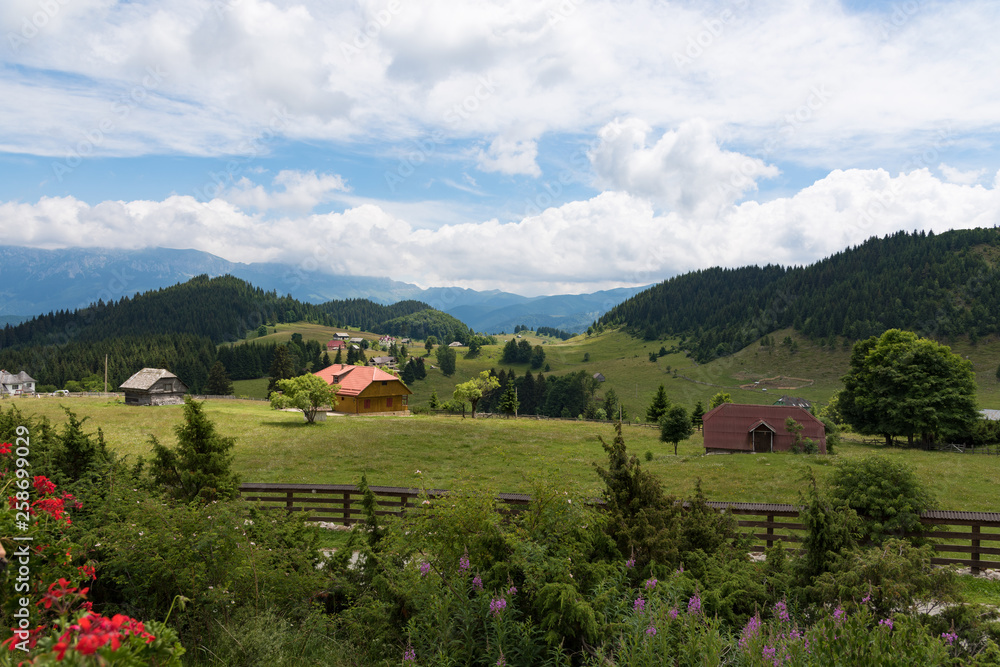 Country view, with cabins and barns at the foot of the mountains. Cloudy blue sky. Lawns and forests