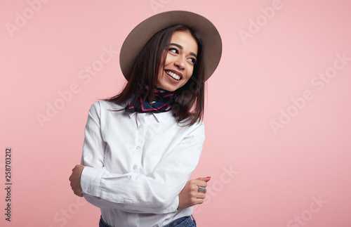 Cheerful pretty lady, laughs happily, expresses sincere emotions, being amused by friend, wearing white shirt and hat. Cute model in studio standing isolated over pink background