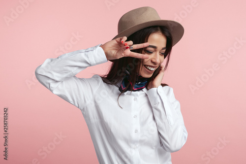 Pleased attractive woman shows v sign or peace gesture, laughs happily, wears white shirt and hat, isolated over rosy studio background. People, fashion, body language.