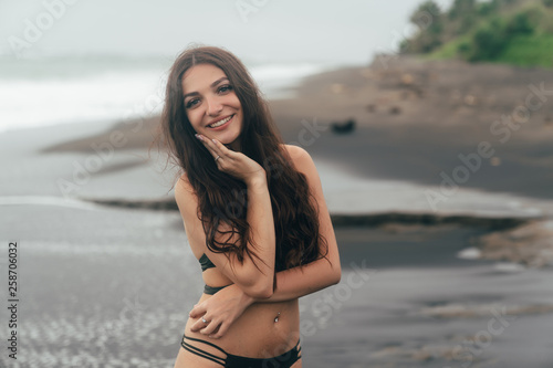 Portrait of tanned smiling girl with closed eyes in swimsuit resting on beach with black sand.