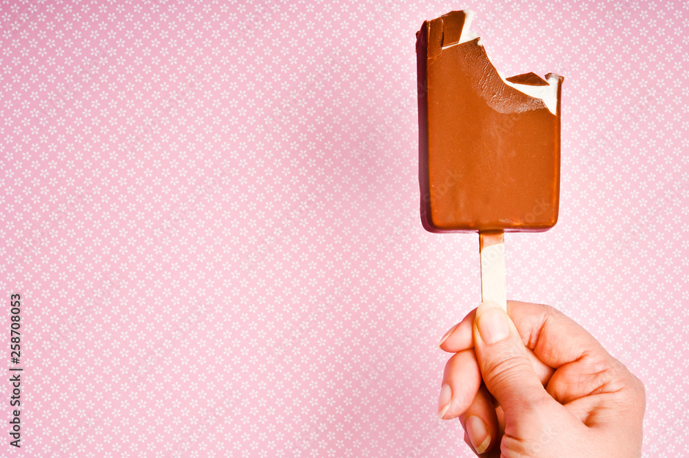 bitten off ice lolly with vanilla icecream and chocolate kept by a female hand over pink background bitten off ice lolly with vanilla icecream and chocolate kept by a female hand over pink background