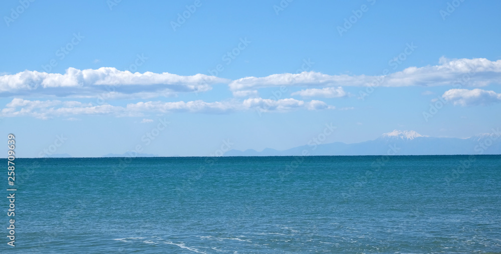Panoramic seascape with sea surface and high mountains silhouettes with white snow cap on top far away on horizon in light haze under blue sky with white cumulus clouds