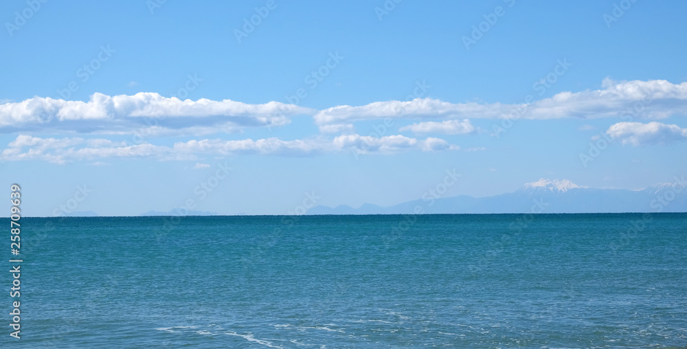 Fototapeta premium Panoramic seascape with sea surface and high mountains silhouettes with white snow cap on top far away on horizon in light haze under blue sky with white cumulus clouds