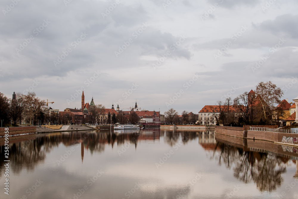 Calm dreamy view of buildings, trees, and gray sky reflected in a crystal clear water of Odra river in Wroclaw, Poland. Typical buildings with red roofs on a background