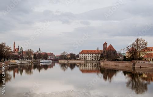 Splendid dreamy view of buildings, trees, and gray sky reflected in a crystal clear water of Odra river in Wroclaw, Poland. Typical buildings with red roofs on a background. Duck swimming in a water