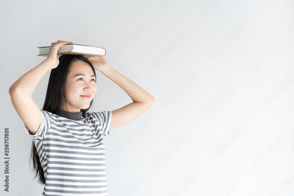 Asian woman student of university standing on a white background holding a book on women head. Women smiling be happy. Female is looking outside Asian woman student of university standing on a white background holding a book on women head. Women smiling be happy. Female is looking outside