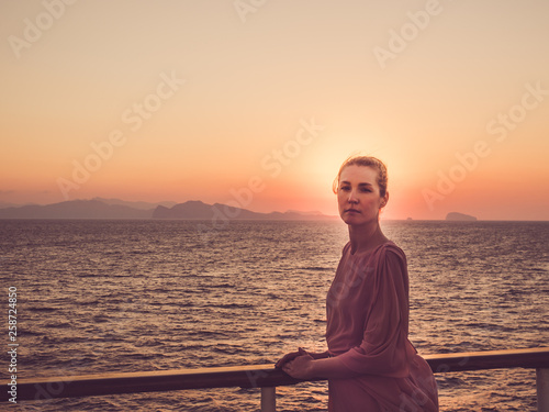 Cute woman in fashionable dress on the top deck of a cruise ship, looking into the distance on a sunset background. Concept of sea cruises and rest