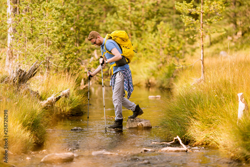 Young man crosses a mountain stream during a hiking on a sunny day