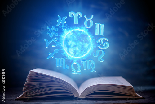 Fototapeta Naklejka Na Ścianę i Meble -  Open book on astrology on a dark background. Glowing magical globe with signs of the zodiac in the blue light.