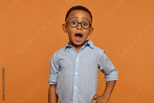 Isolated picture of handsome emotional bug eyed dark skinned schoolboy wearing stylish glasses expressing shock, keeping mouth wide opened. Excitement, fascination and true genuine emotions photo