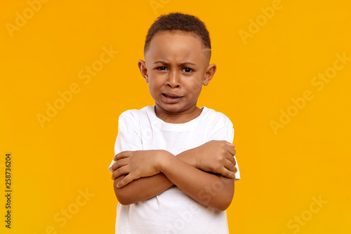 Photography Portrait of stubborn reluctant black dark skinny child posing in studio with grumpy dissatisfied grimace, keeping arms on his chest, demonstrating unwillingness to clean his room