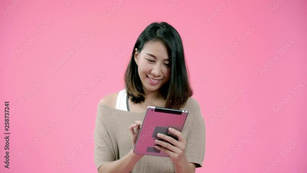 Young Asian woman using tablet checking social media feeling happy smiling in casual clothing over pink background studio shot. Happy smiling adorable glad woman rejoices success. Young Asian woman using tablet checking social media feeling happy smiling in casual clothing over pink background studio shot. Happy smiling adorable glad woman rejoices success.
