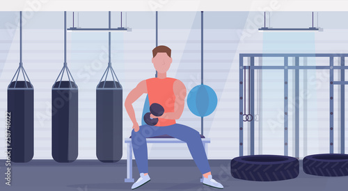 sports man doing exercises with dumbbell muscular guy sitting on bench biceps workout concept training in gym with punching bags modern health club interior flat full length