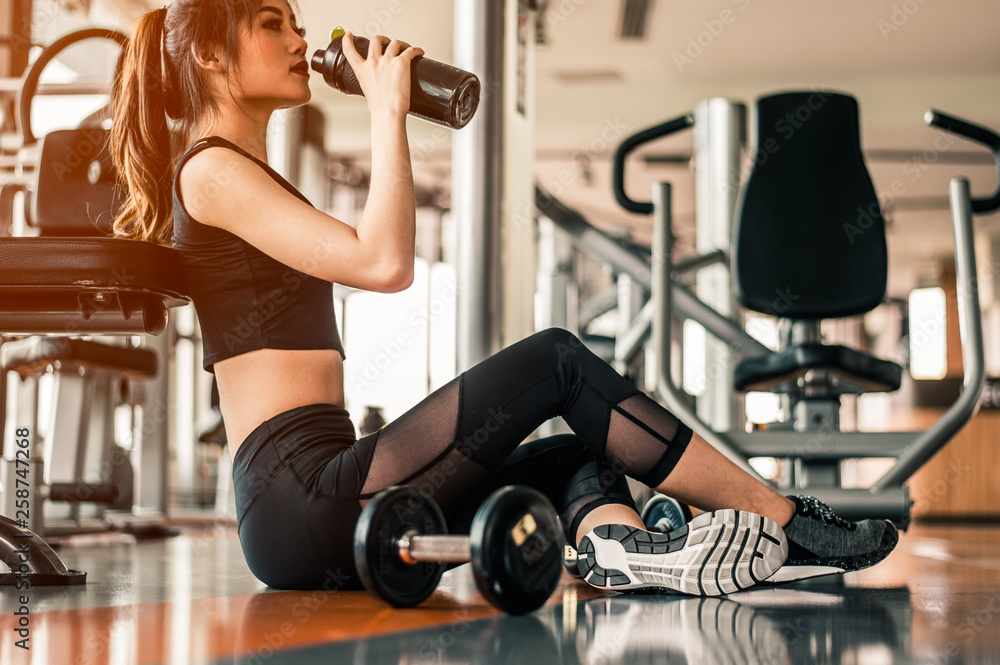 Fitness woman Relaxing after exercise with a whey protein and dumbbell placed beside the gym.Relaxing after training.beautiful young woman looking away while sitting at gym. Fitness woman Relaxing after exercise with a whey protein and dumbbell placed beside the gym.Relaxing after training.beautiful young woman looking away while sitting at gym.