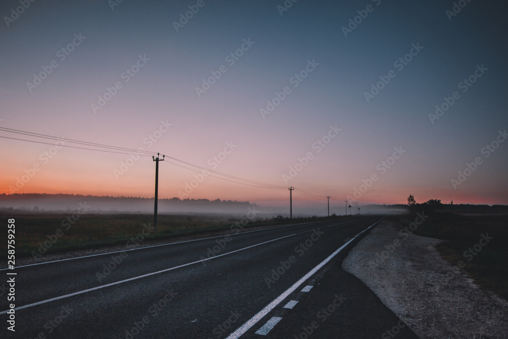 Morning has come. The orange sun rises above a misty field, asphalt road and forest, dark trees. Morning has come. The orange sun rises above a misty field, asphalt road and forest, dark trees.