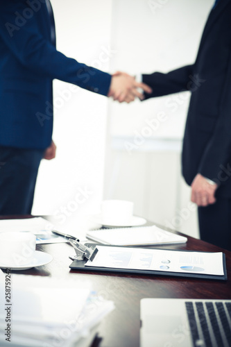 Documents and laptop on the table. Business people shaking hands on the background, silhouettes