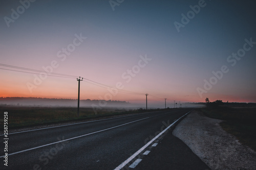Morning has come. The orange sun rises above a misty field, asphalt road and forest, dark trees. photo