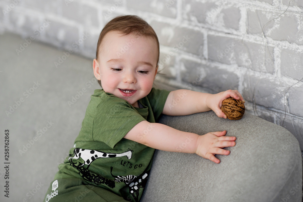 Naklejka premium The one-year-old boy is sitting on a gray sofa with a walnut and smiled happily. Positive lifestyle concept. Happy childhood.