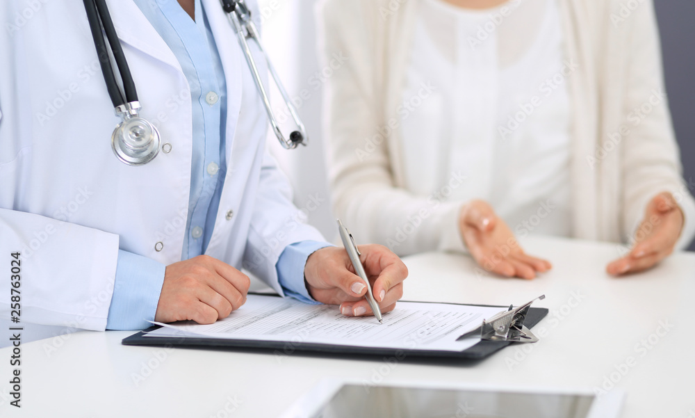 Unknown woman doctor and female patient discussing something while standing near reception desk in emergency hospital. Physician at work in clinic. Medicine and health care concept Unknown woman doctor and female patient discussing something while standing near reception desk in emergency hospital. Physician at work in clinic. Medicine and health care concept