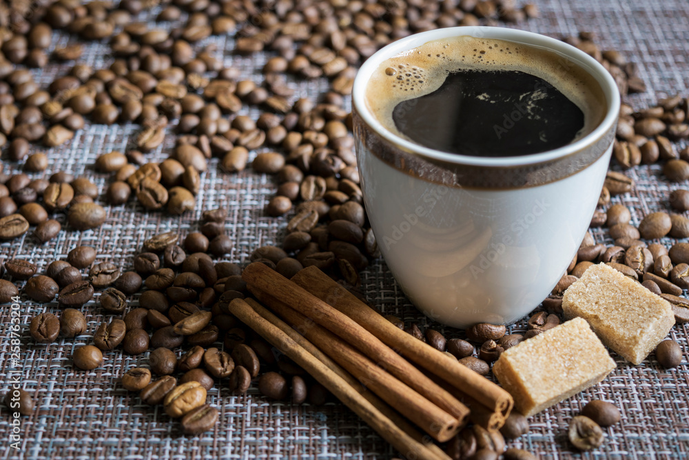 A Cup of coffee with foam stands on the table among the scattered coffee beans. Next to the Cup are pieces of sugar and cinnamon sticks.