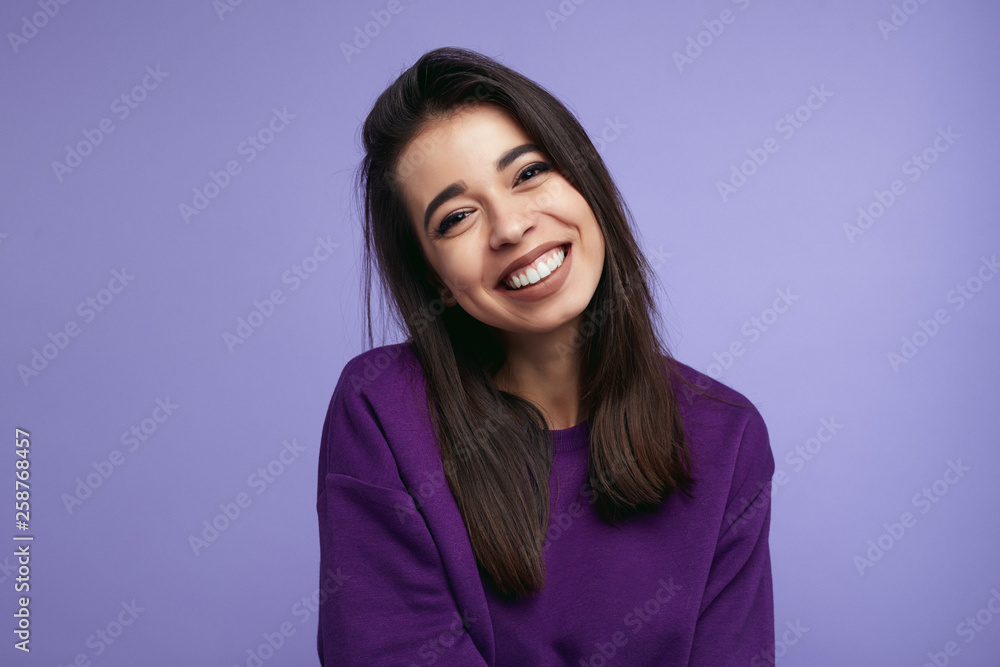 Portrait of pretty adult girl, broad smile, wears purple sweatshirt, tilts head and looks with joy, dressed casually, isolated over violet background Portrait of pretty adult girl, broad smile, wears purple sweatshirt, tilts head and looks with joy, dressed casually, isolated over violet background